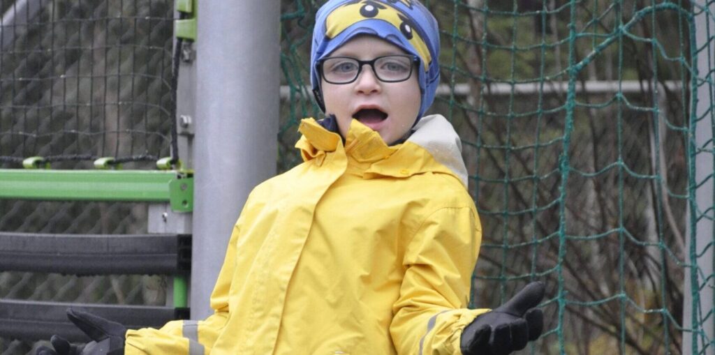Foto av en gutt med Noonans syndrom med briller, gul jakke og blå lue som leker ute