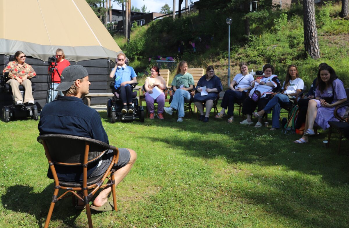 En gruppe deltaker sitter i en halvmåne foran en mann som sitter i en stol, og blir spurt forskjellige spørsmål.