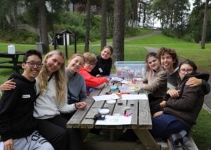 En gjeng med mennesker sitter rundt et bord på gressletta. De smiler og klemmer hverandre.