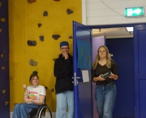 To mennesker gjemmer seg bak en dør, som en tredje person er på vei til å gå inn gjennom. De bak døren skal skremme den som går inn.